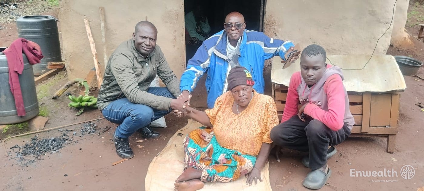 An older woman seated on a mat outside a mud-walled house, a young man beside her, two Foundation staff shaking her hand.
